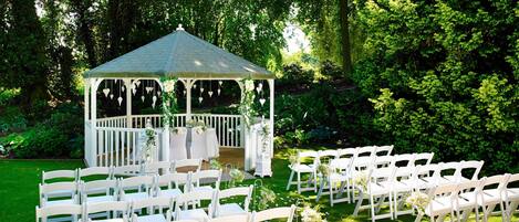 A gazebo with white chairs arranged in rows, surrounded by lush greenery.