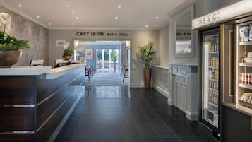Una hall d'albergo con una reception, un cartello con la scritta 'CAST IRON BAR & GRILL', un refrigeratore con prodotti 'GRAB & GO' e una grande pianta in vaso.
