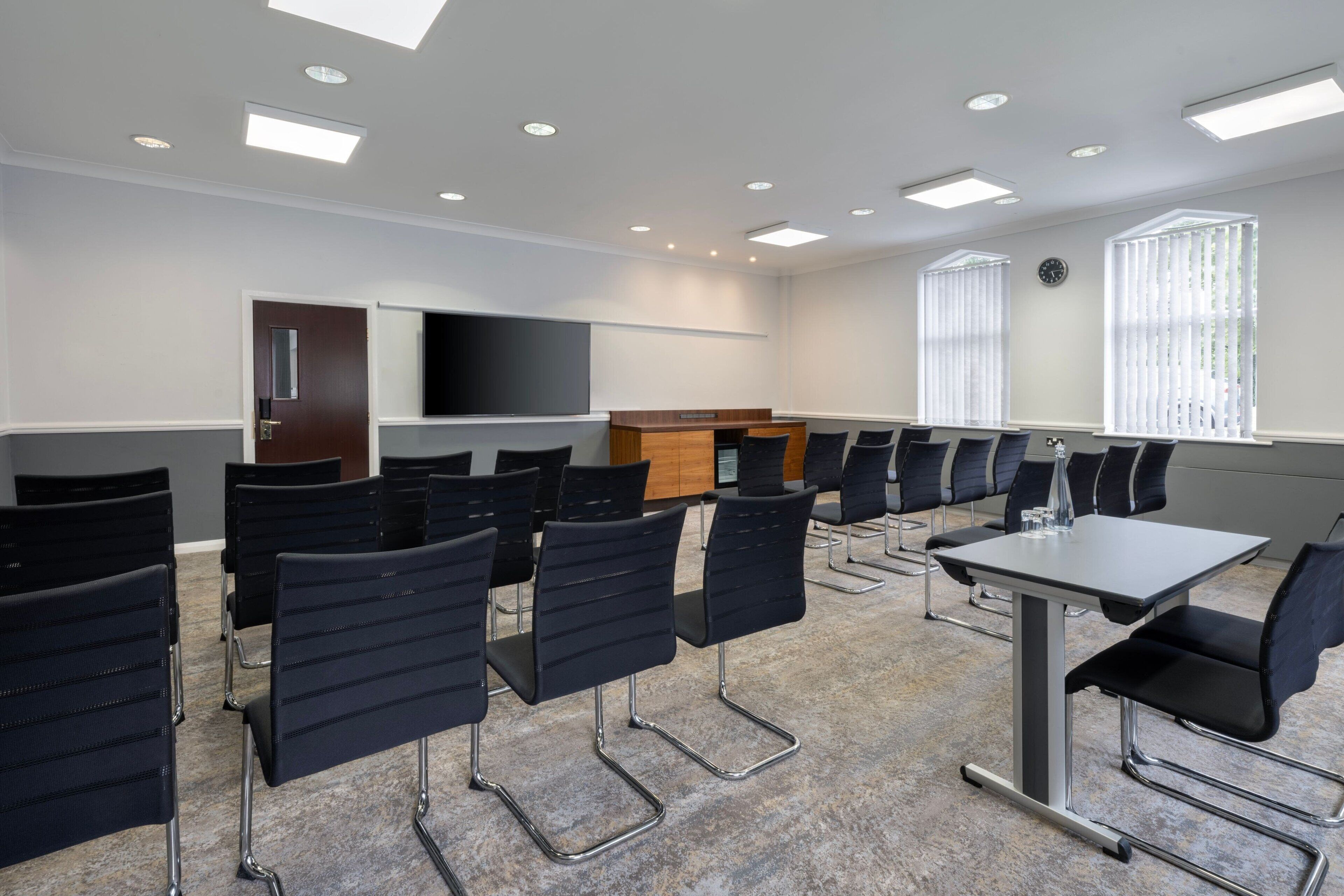 A conference room with a large flat-screen TV, a wooden podium, and rows of black chairs.