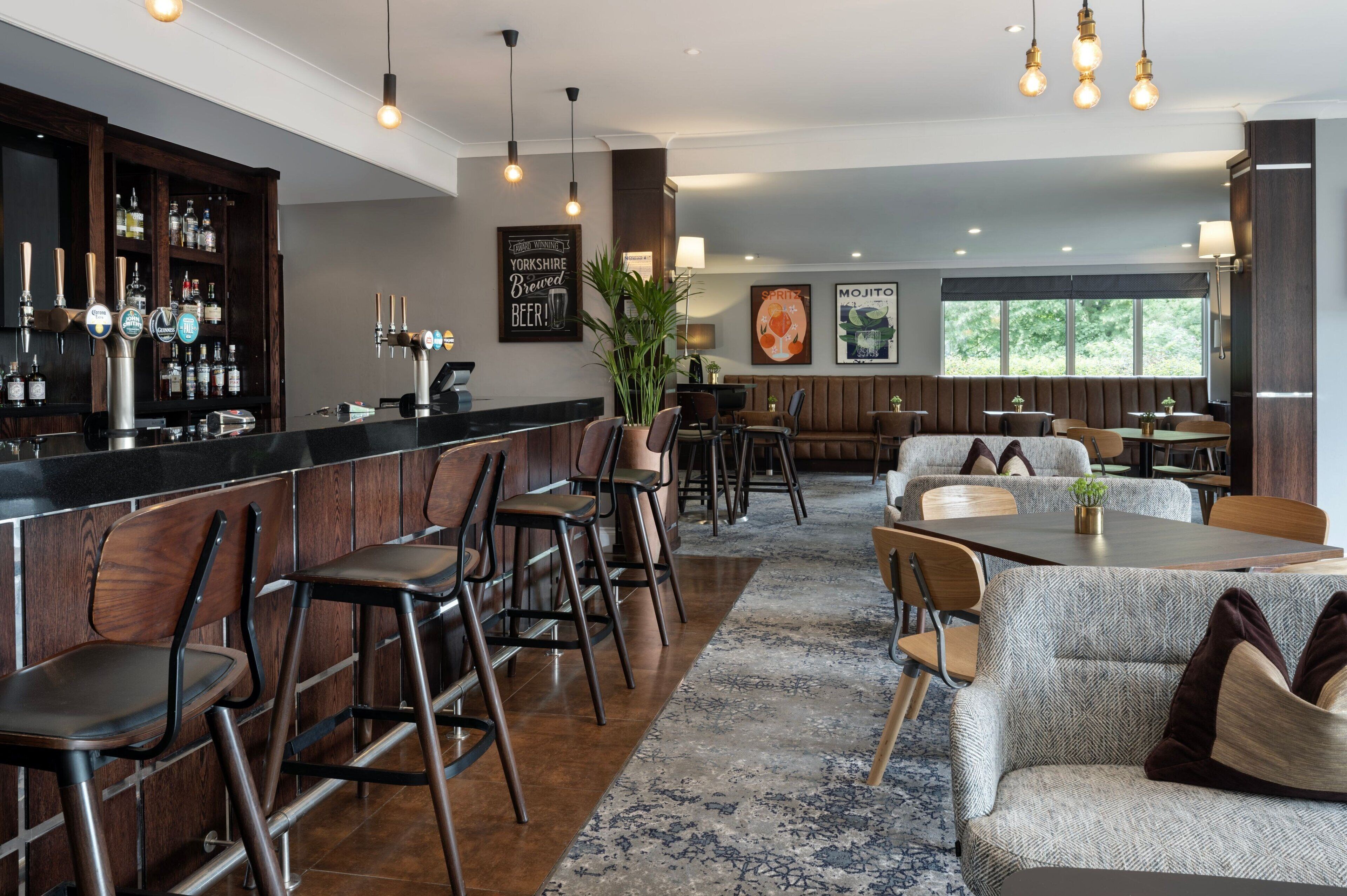 A bar area with stools and a counter, a seating area with tables and chairs, and a large window with a view of greenery.