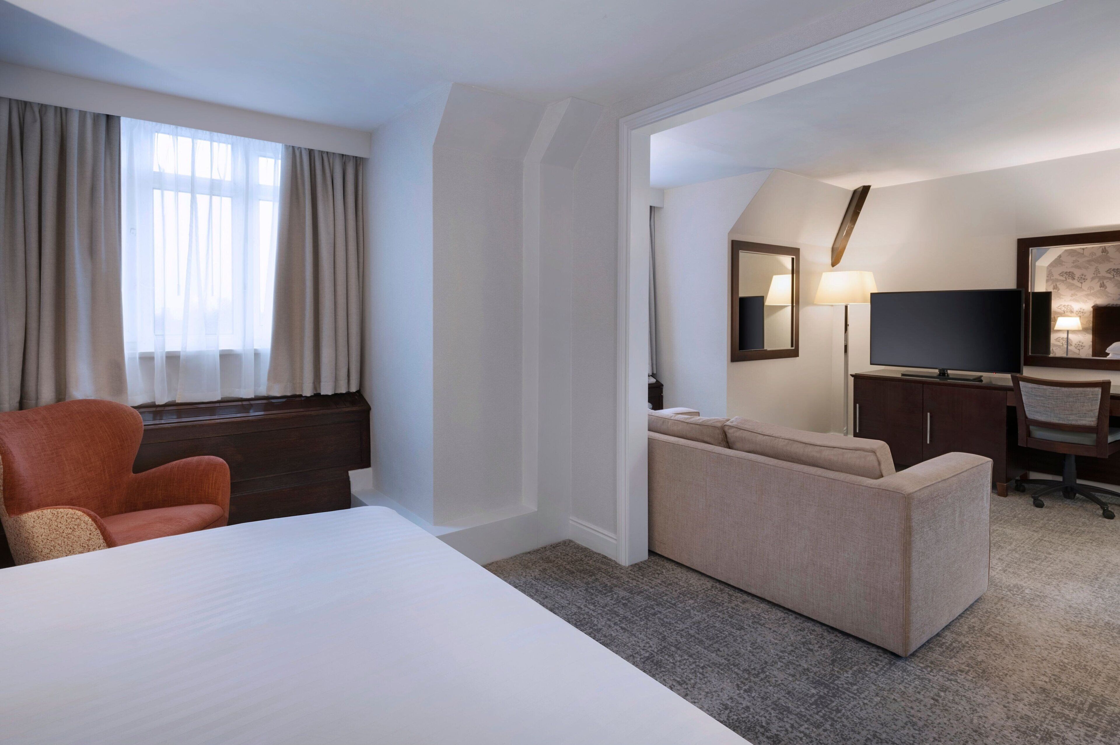A hotel room with a bed, a desk, a chair, a sofa, a TV, and a window with curtains.