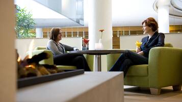 Two people seated on green sofas in a modern indoor setting with a glass partition, a small table with a drink, and a vase with a flower.
