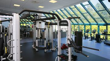 Et fitnesscenter med forskellig træningsudstyr, et stort ovenlys og udsigt til træer udefra.