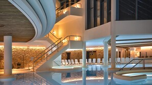 A modern indoor swimming pool with a curved ceiling, multiple levels, and seating areas.