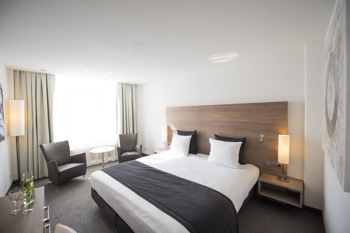 Habitació d'hotel amb un llit gran, una cadira, una taula petita i una làmpada.