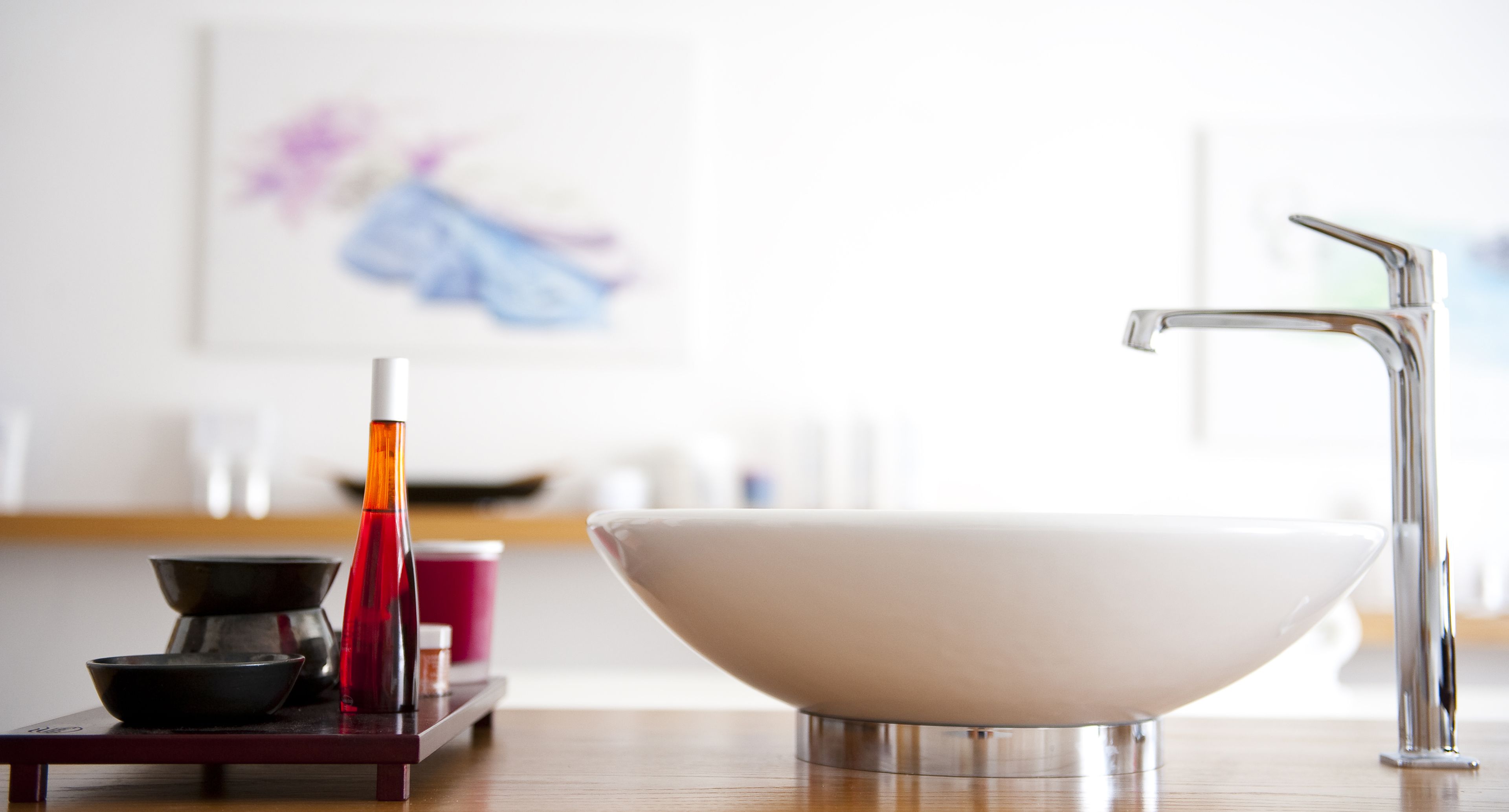A modern bathroom with a white sink, a silver faucet, and a wooden tray holding a black bowl, a red bottle, and a small container.