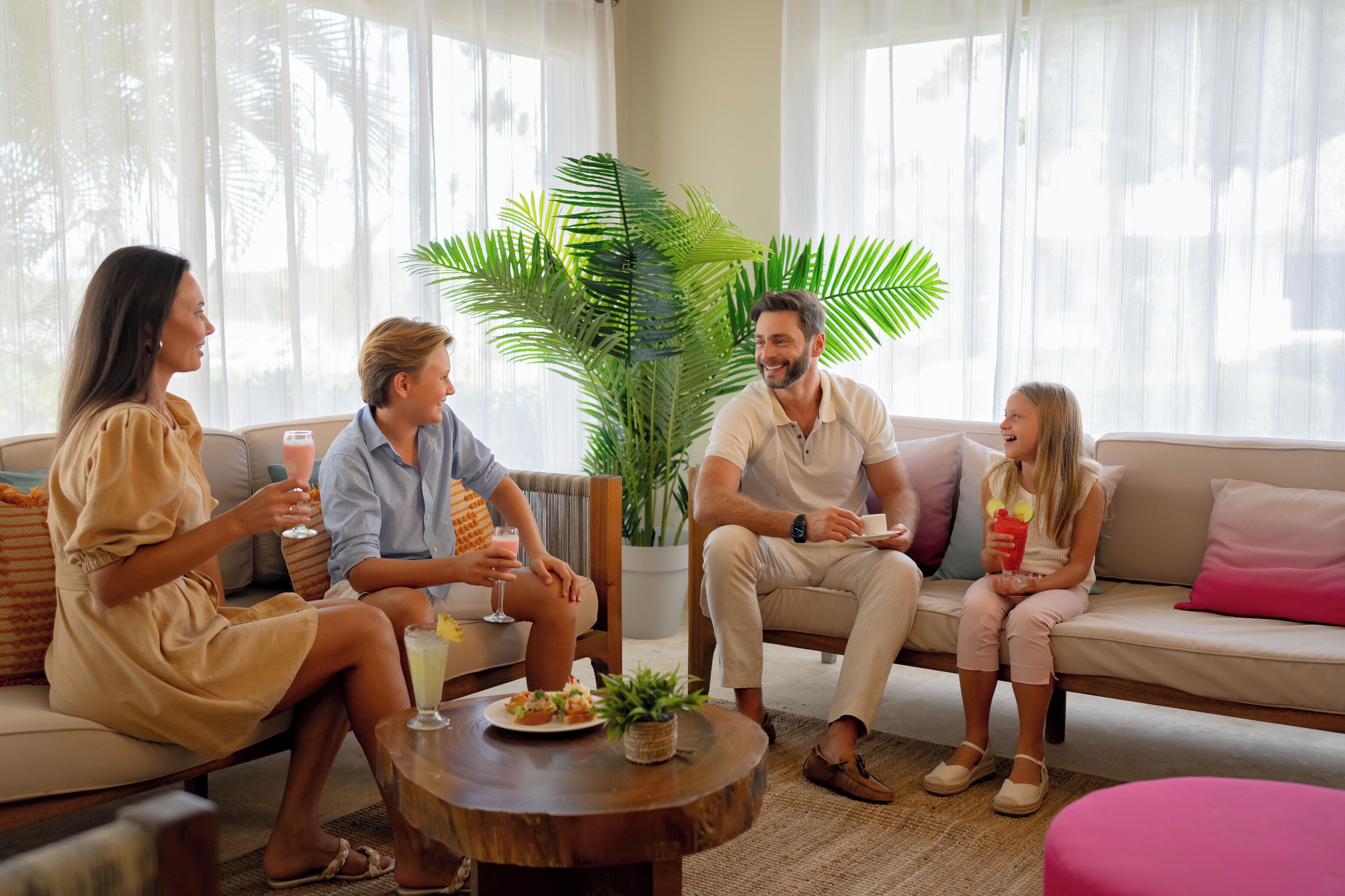 A family of four seated on a sofa in a well-lit room with a large plant and a coffee table.
