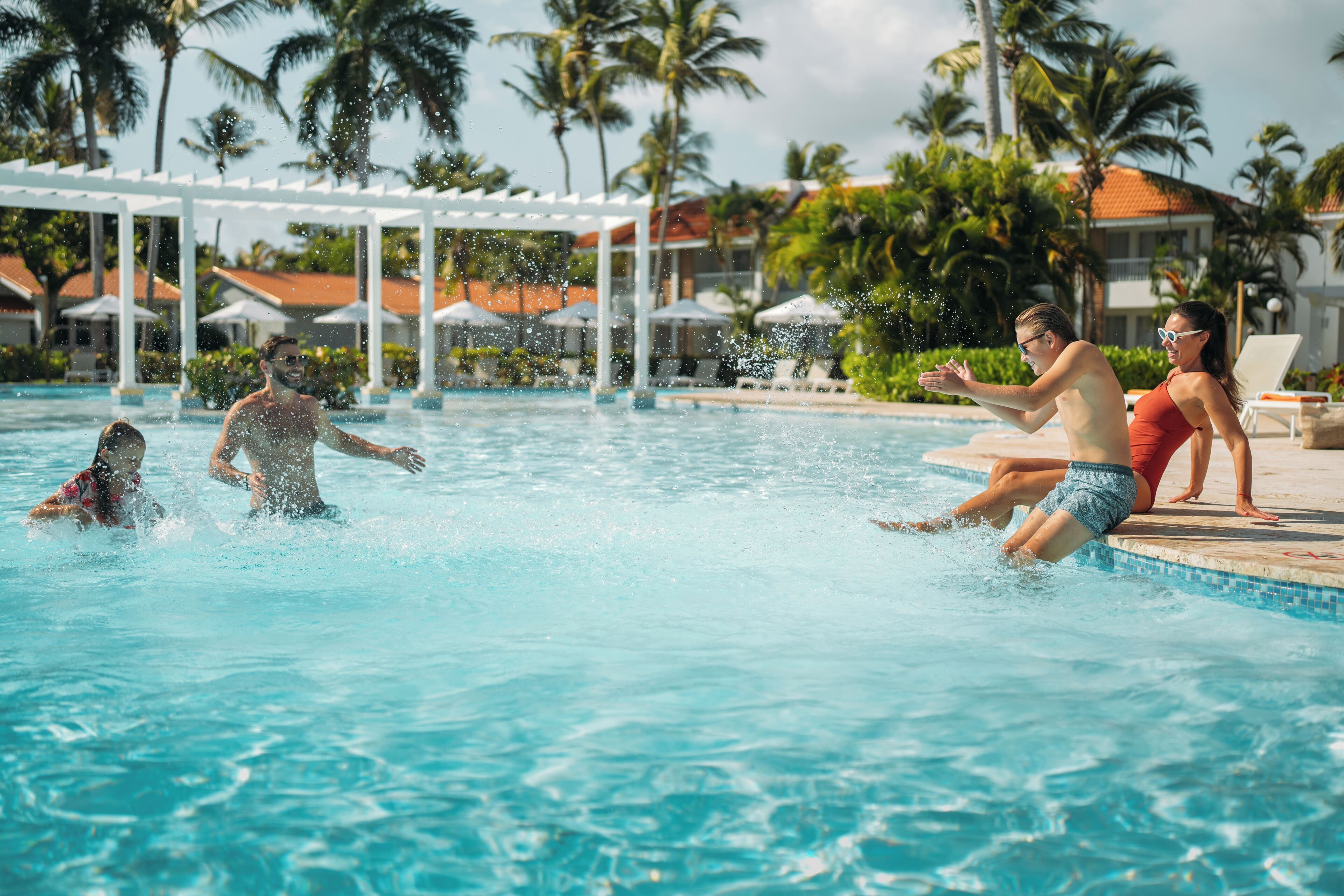 A swimming pool with people in the water, surrounded by palm trees and a covered area with umbrellas.