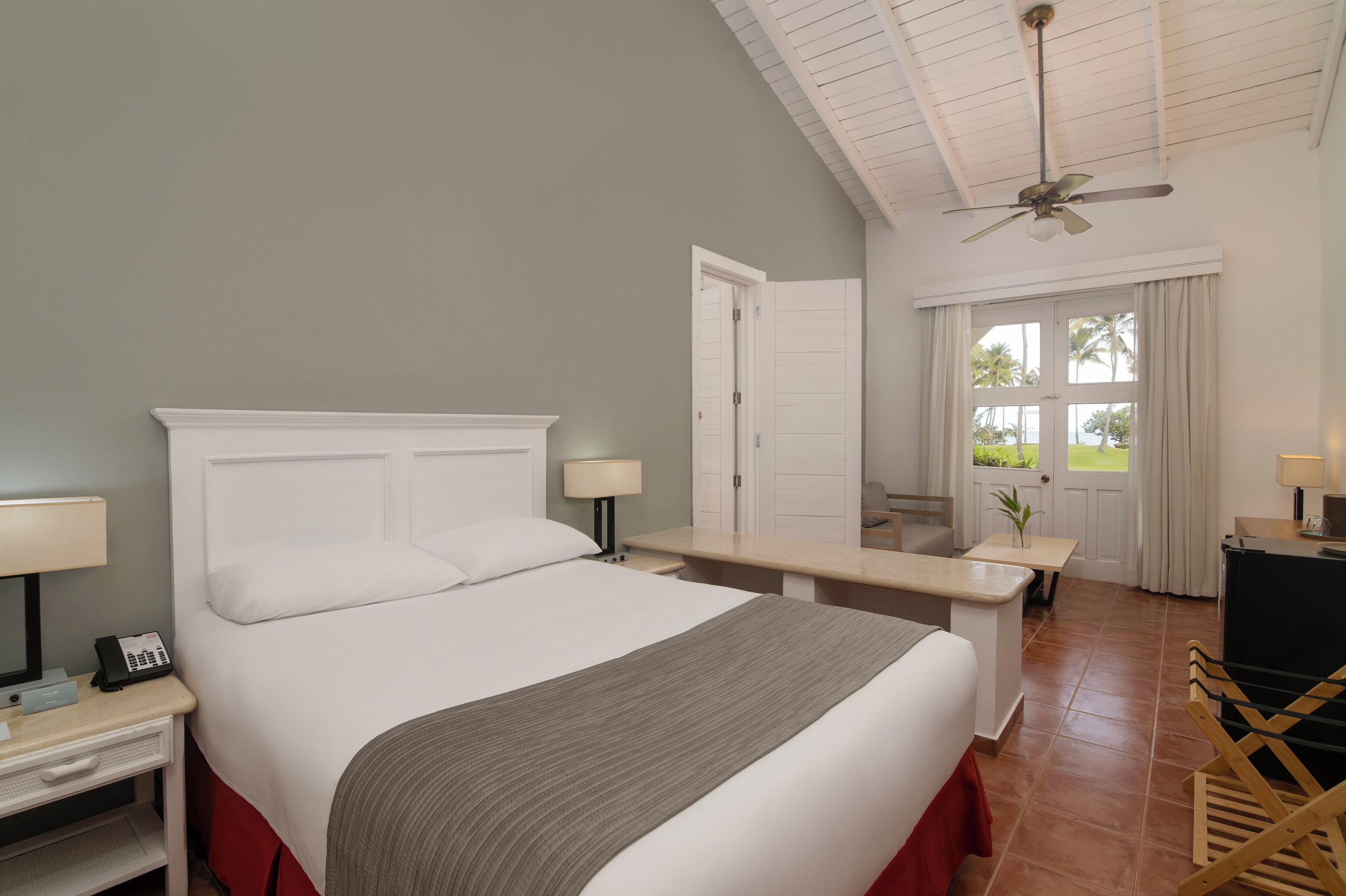 A hotel room with a large bed, bedside tables, a desk, a ceiling fan, and a view of the outdoors through a door.