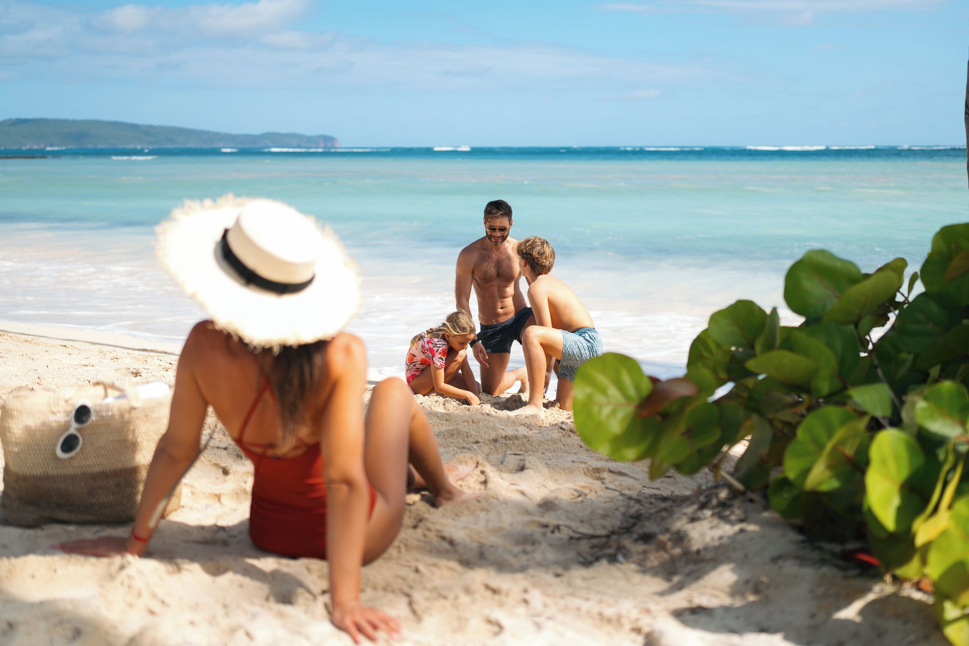 A beach scene with people sitting on the sand, a straw hat, and a beach bag.