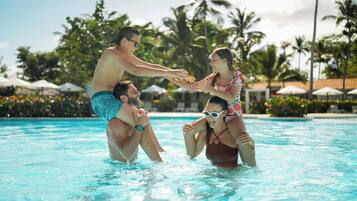 Área de piscina com pessoas brincando na água, cercada por palmeiras e guarda-sóis.