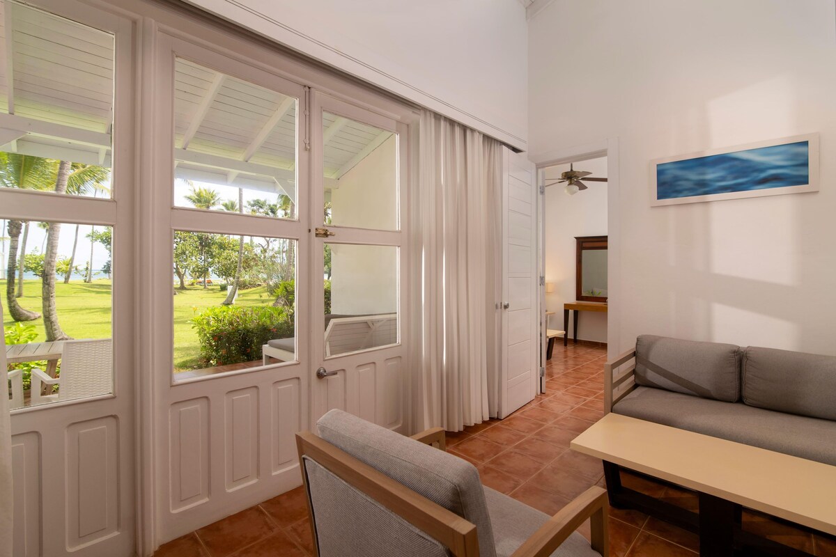 A room with a sliding glass door leading to a garden, a sofa, a chair, a table, and a ceiling fan.