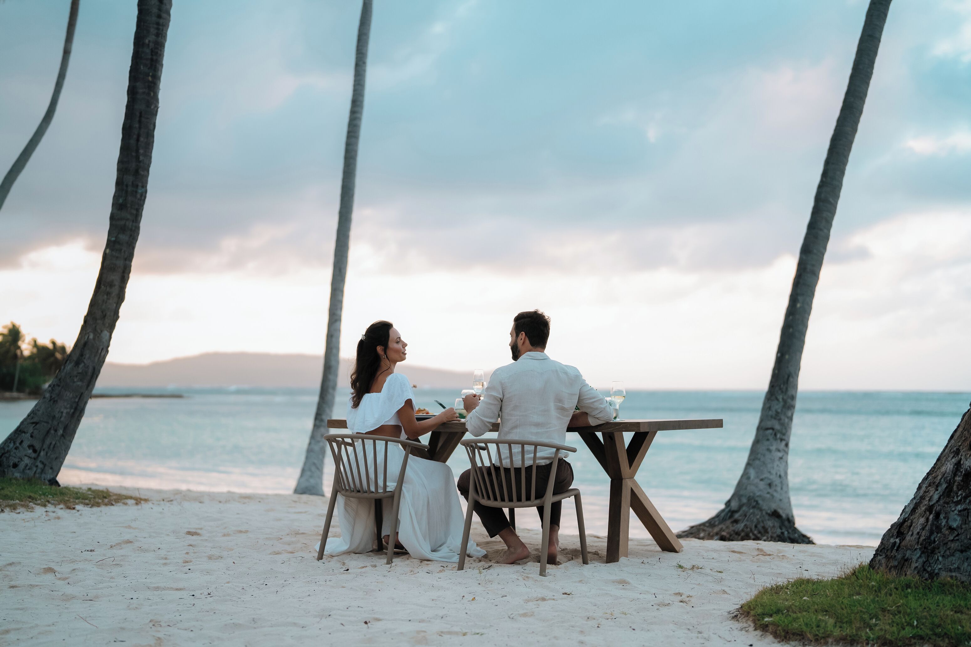 A couple dining at a wooden table on a sandy beach with palm trees and a view of the ocean.