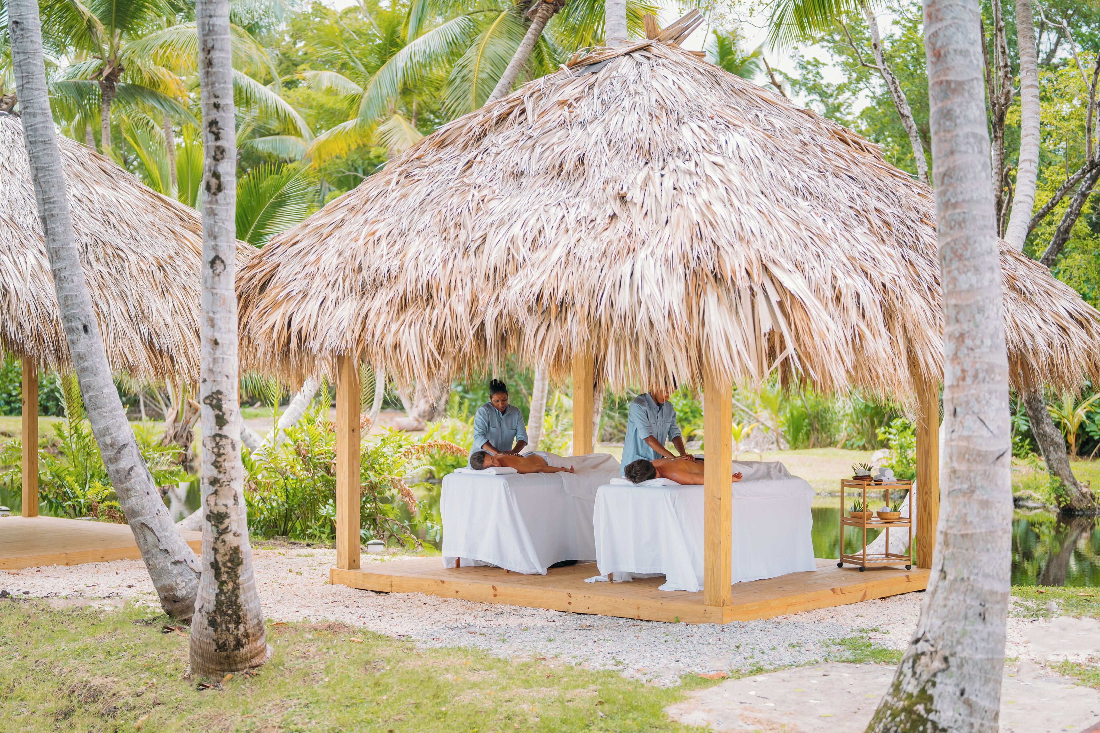 A thatched-roof structure with two massage tables, surrounded by palm trees and greenery.