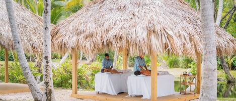 A thatched-roof structure with two massage tables, surrounded by palm trees and greenery.