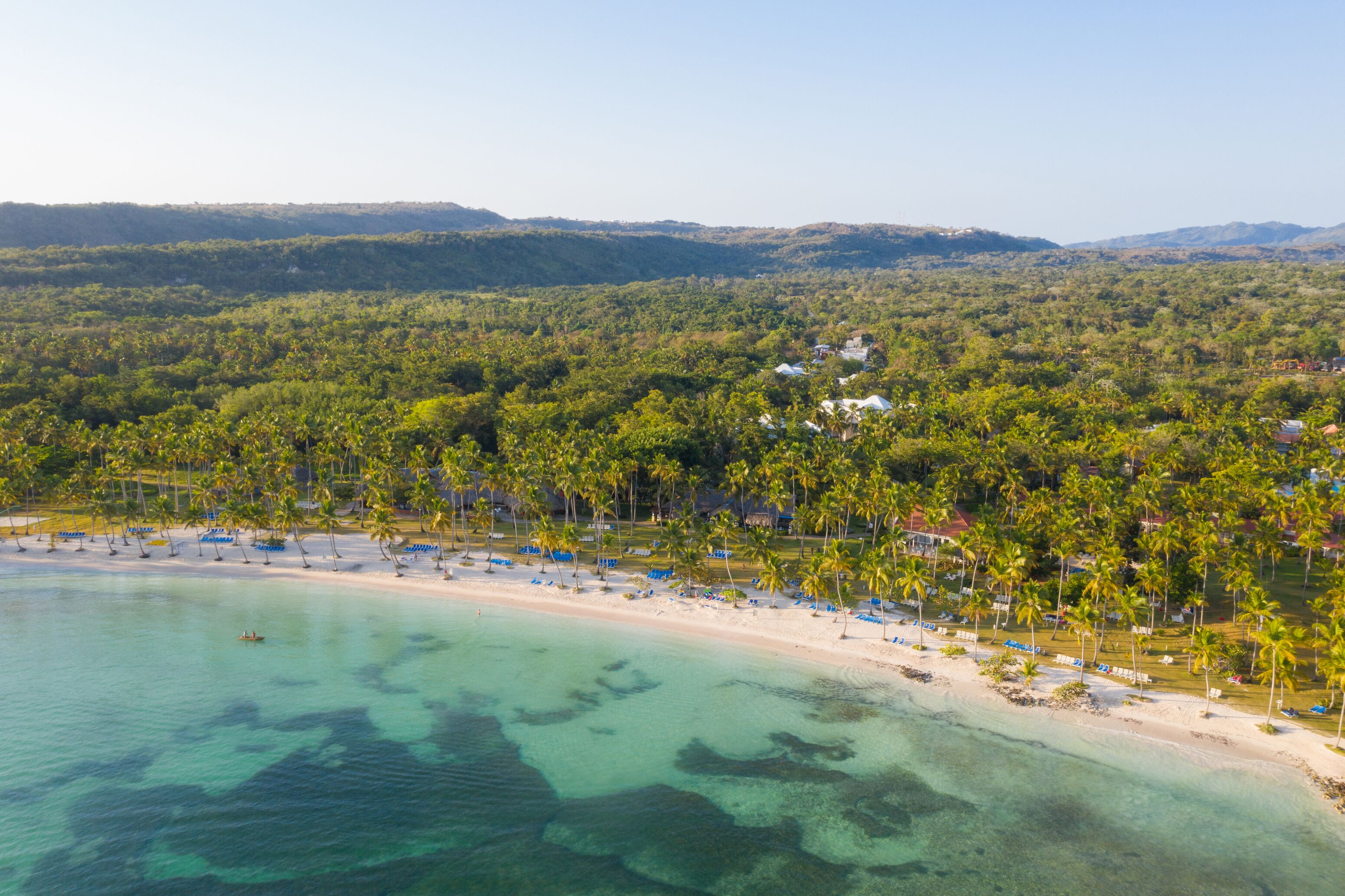 A beach resort with clear turquoise waters, palm trees, and a hilly backdrop.