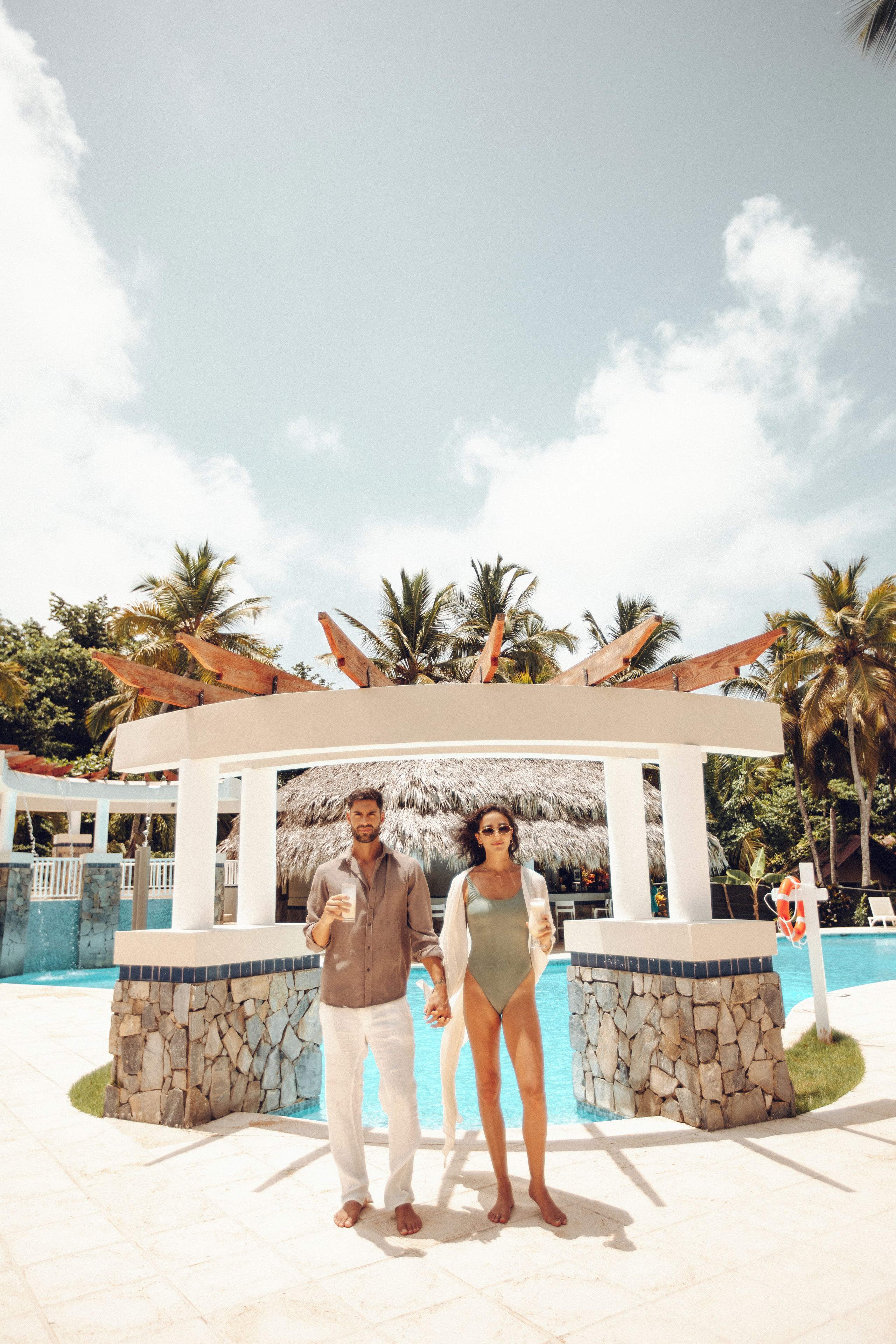 A couple standing in front of a pool with a thatched roof structure, palm trees, and a clear sky.