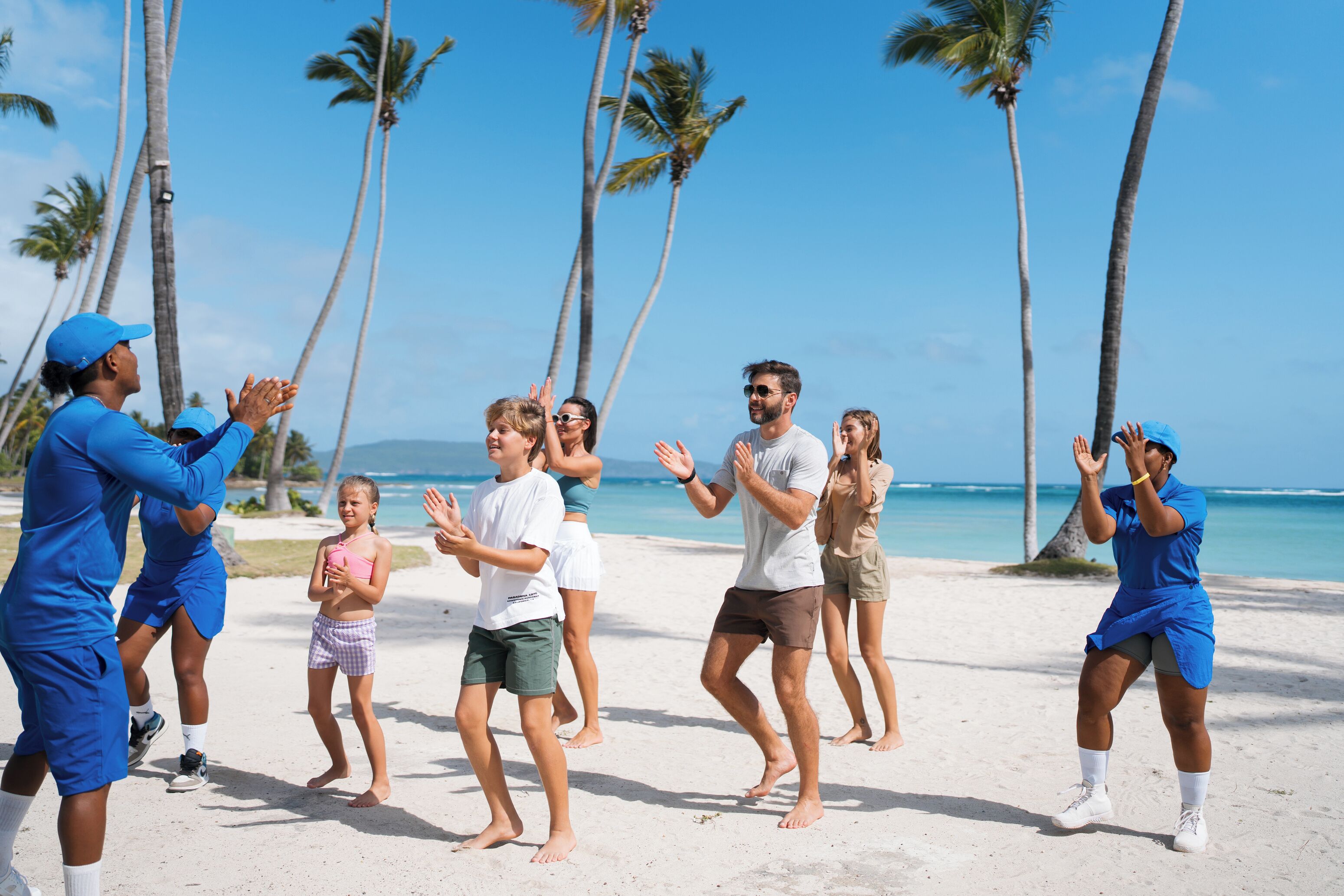 Un grupo de personas realizando una danza sincronizada en una playa con palmeras y el océano al fondo.