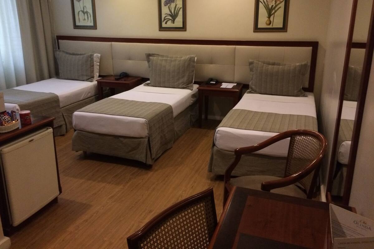 A hotel room with two single beds, a small table, a chair, a desk, and a refrigerator.