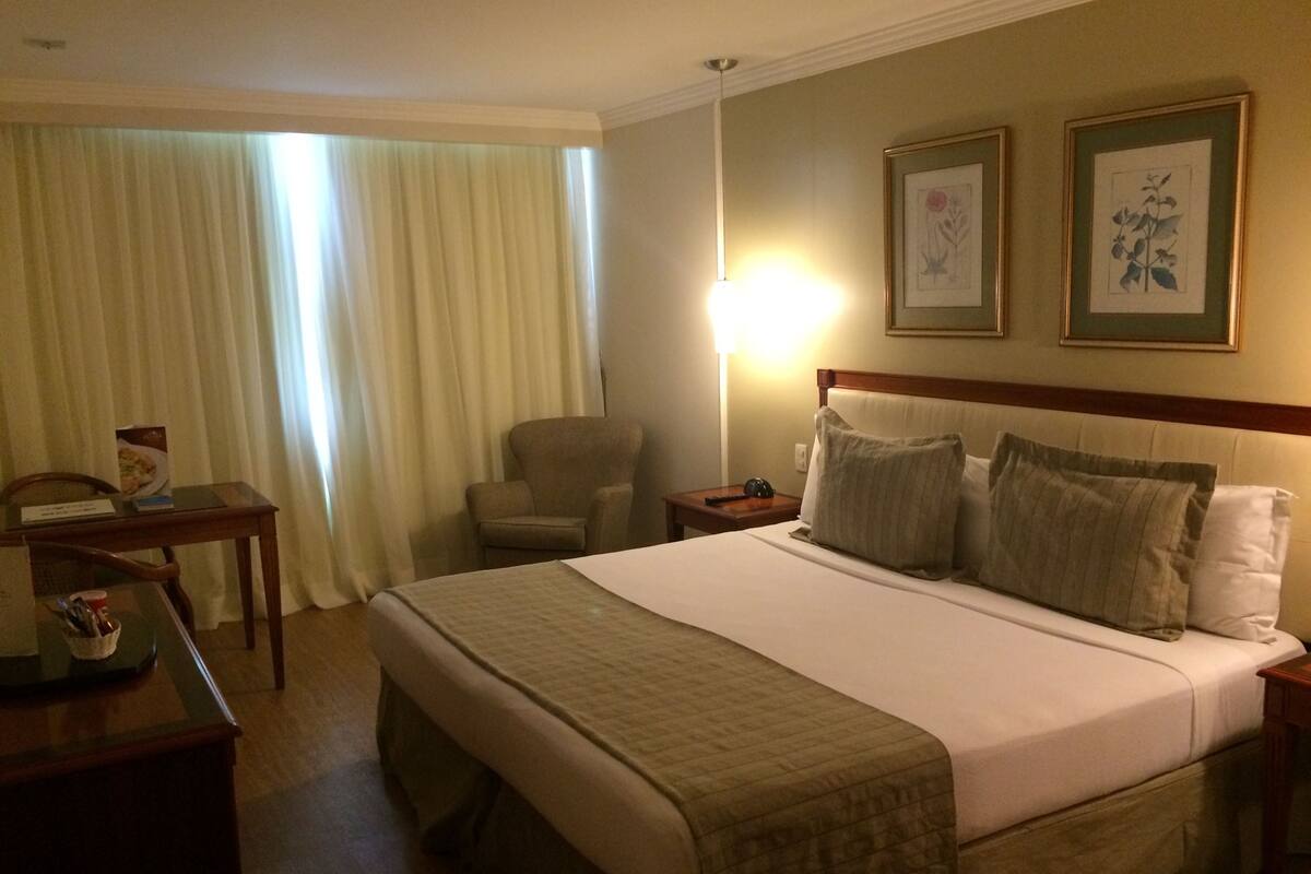 A hotel room with a large bed, a desk with a chair, a lamp, and two framed botanical prints on the wall.