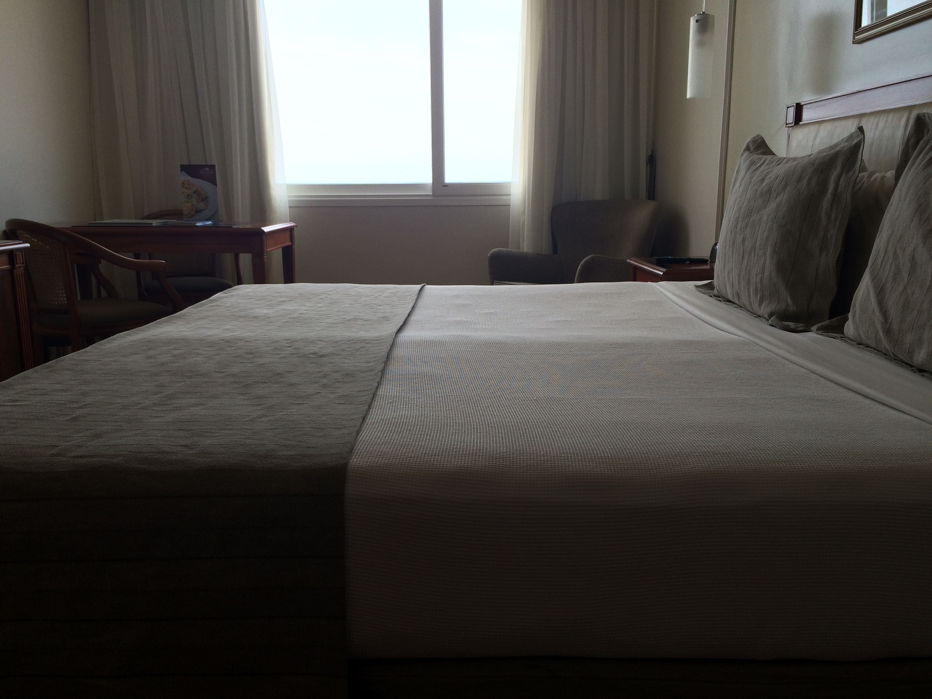 A neatly made bed with a textured blanket, a wooden nightstand, a chair, and a desk with a lamp.