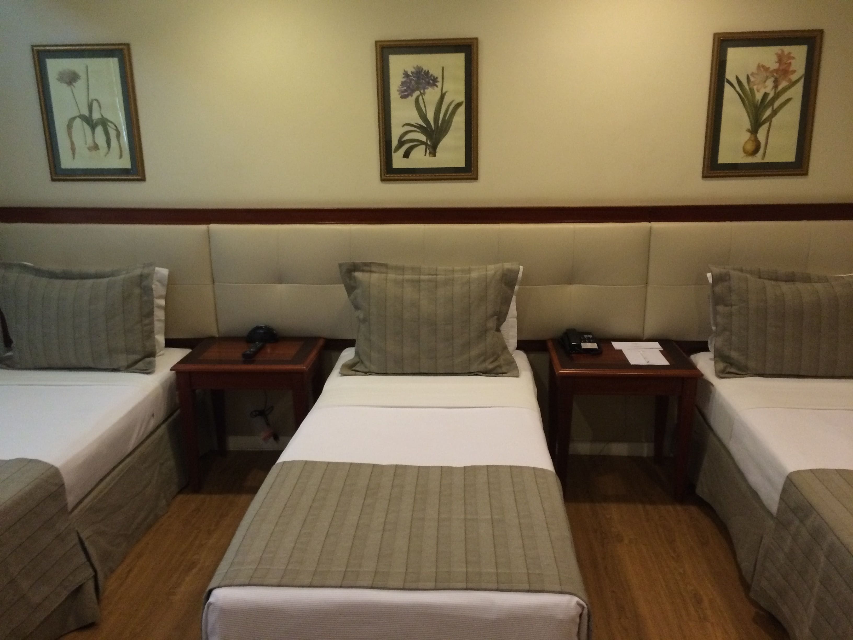 A hotel room with two single beds, a central bed, and a wooden headboard. There are framed floral pictures on the wall.