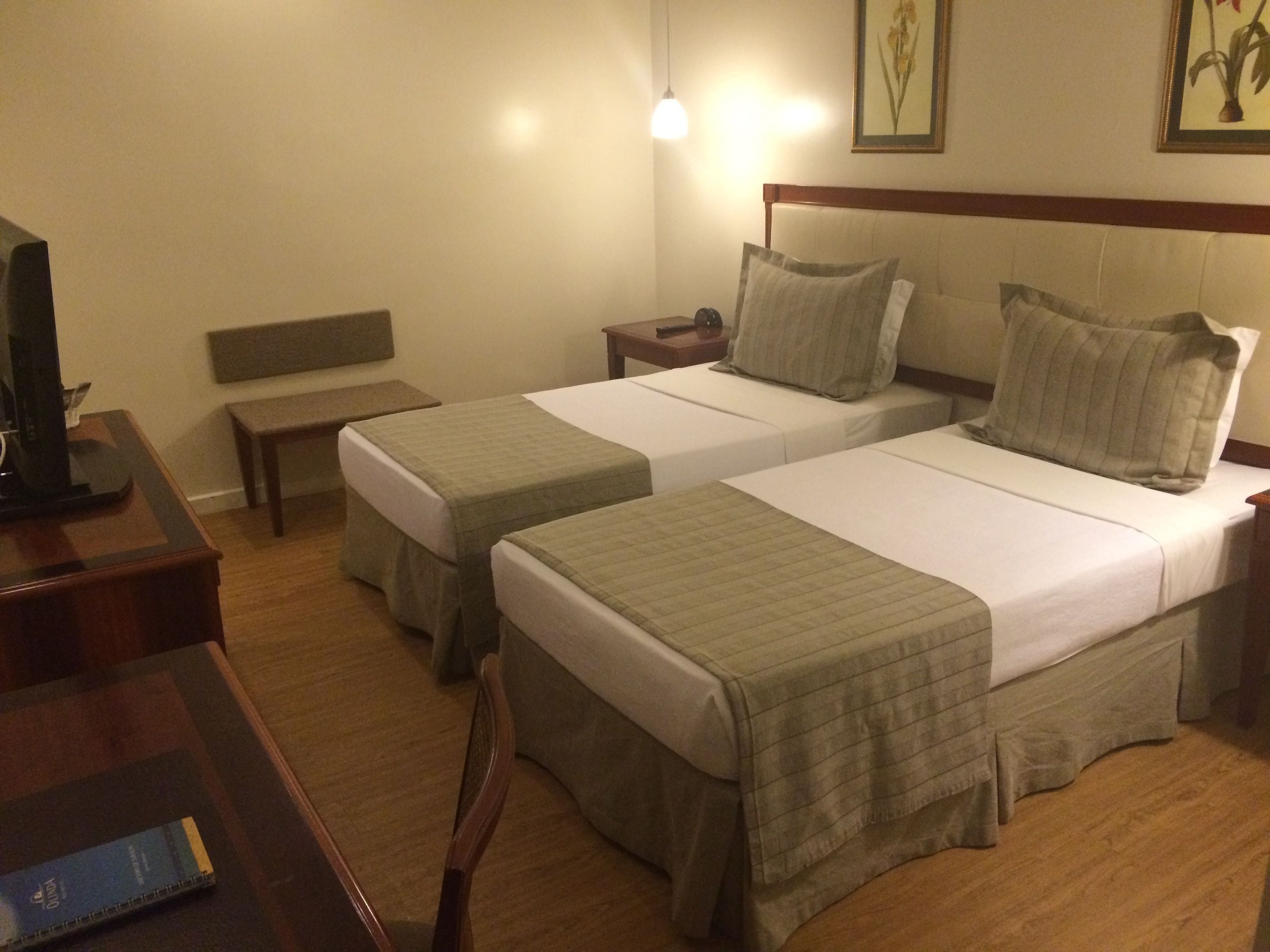 Une chambre d’hôtel avec deux lits simples, une table de chevet avec une lampe, un banc, une télévision et un tableau encadré au mur.