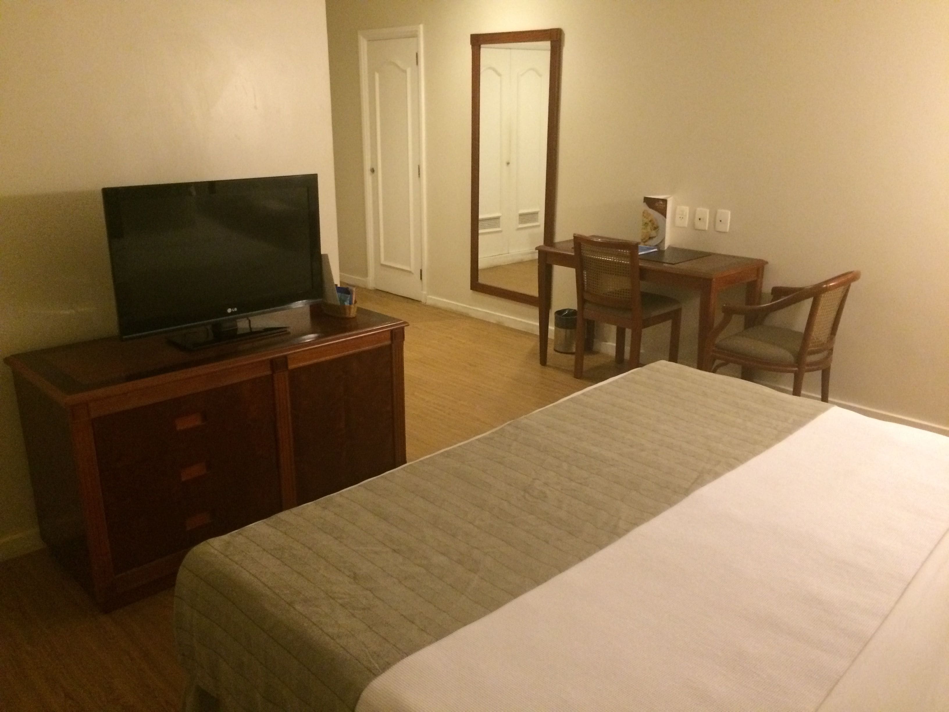 A hotel room with a bed, a TV on a wooden dresser, a desk with a chair, and a mirror.