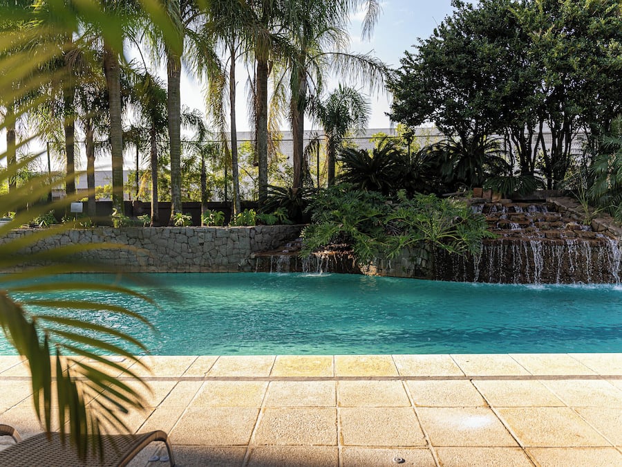 Piscina con caída de agua rodeada de follaje tropical y una pared de piedra.