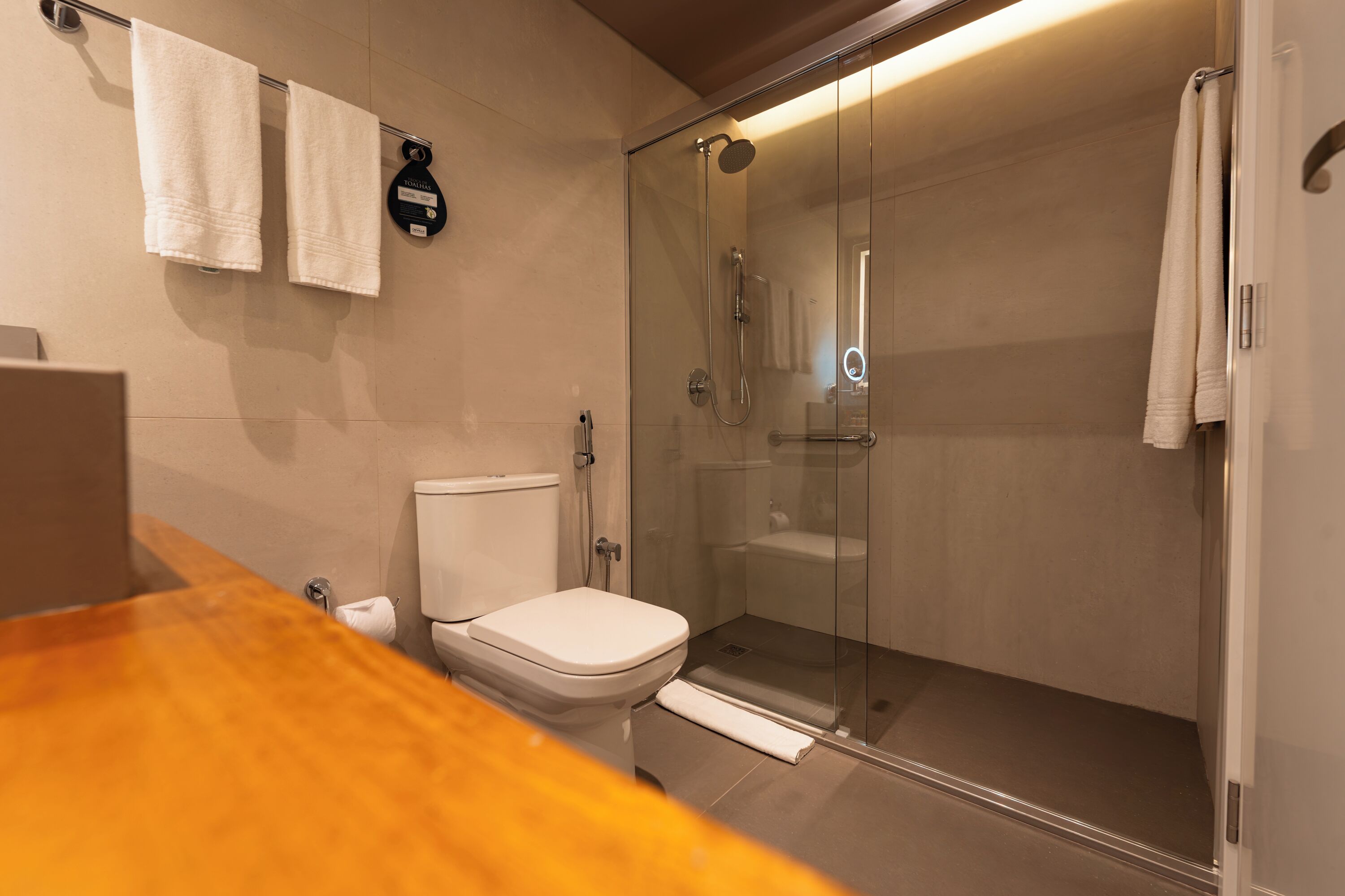 Uma casa de banho moderna com chuveiro em cabine de vidro, sanita e lavatório com bancada de madeira.