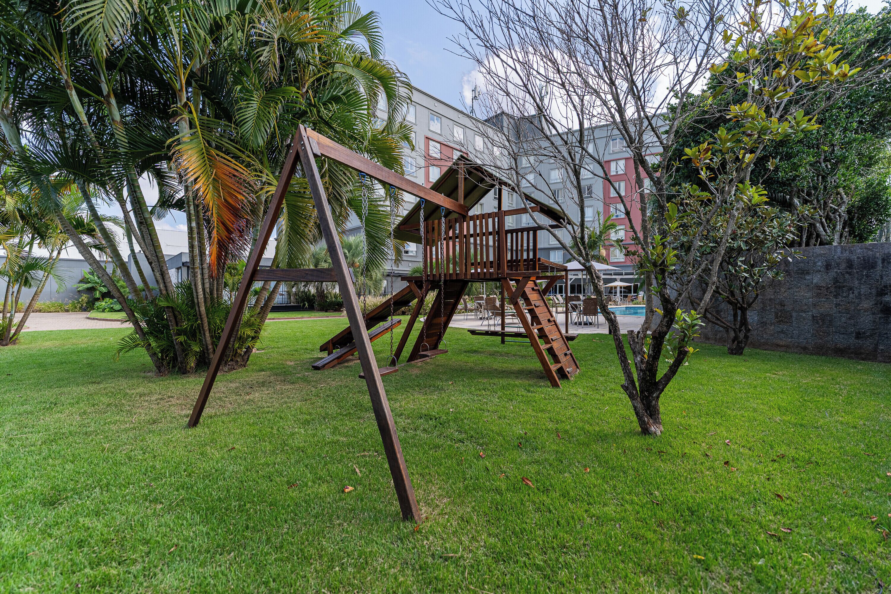 Un parque infantil con columpios y una estructura para escalar en un área verde con árboles y un edificio al fondo.