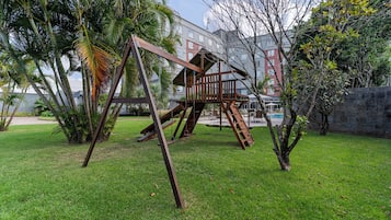 Un parque infantil con columpios y una estructura para escalar en un área verde con árboles y un edificio al fondo.