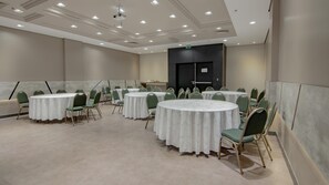 Un salón de conferencias con varias mesas redondas y sillas, un proyector y una puerta señalizada con 'SAIDA'.