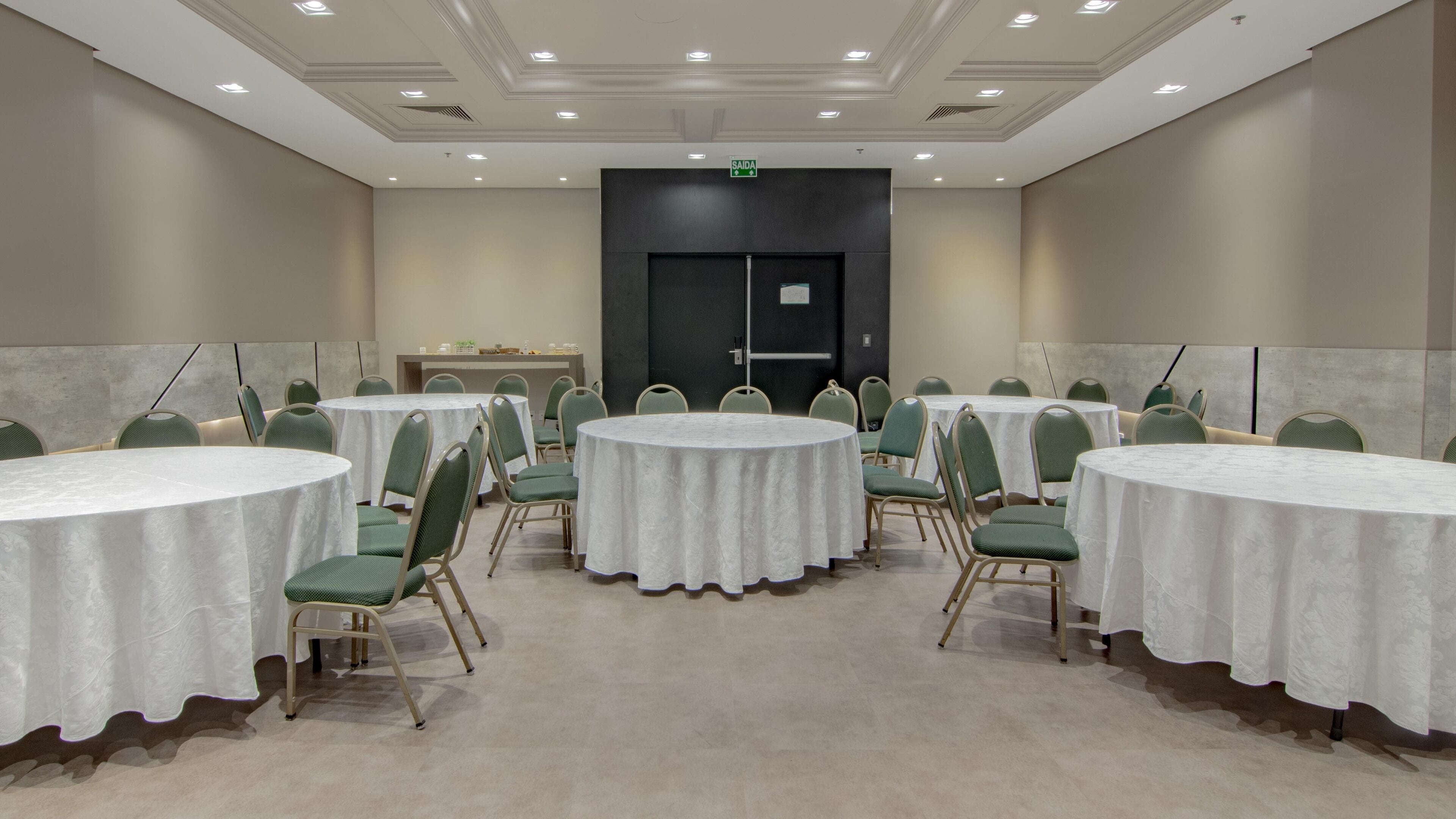 Un salón de conferencias con mesas redondas y sillas verdes, una puerta con un letrero y un techo con iluminación empotrada.