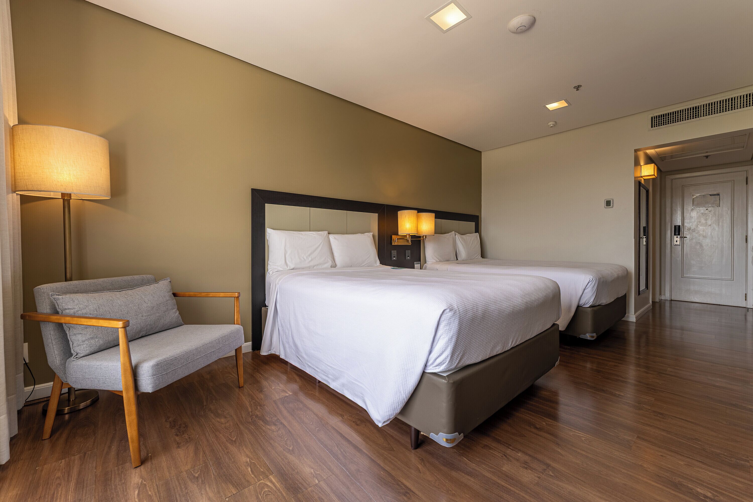 Quarto de hotel com cama, abajures, uma cadeira e uma porta.