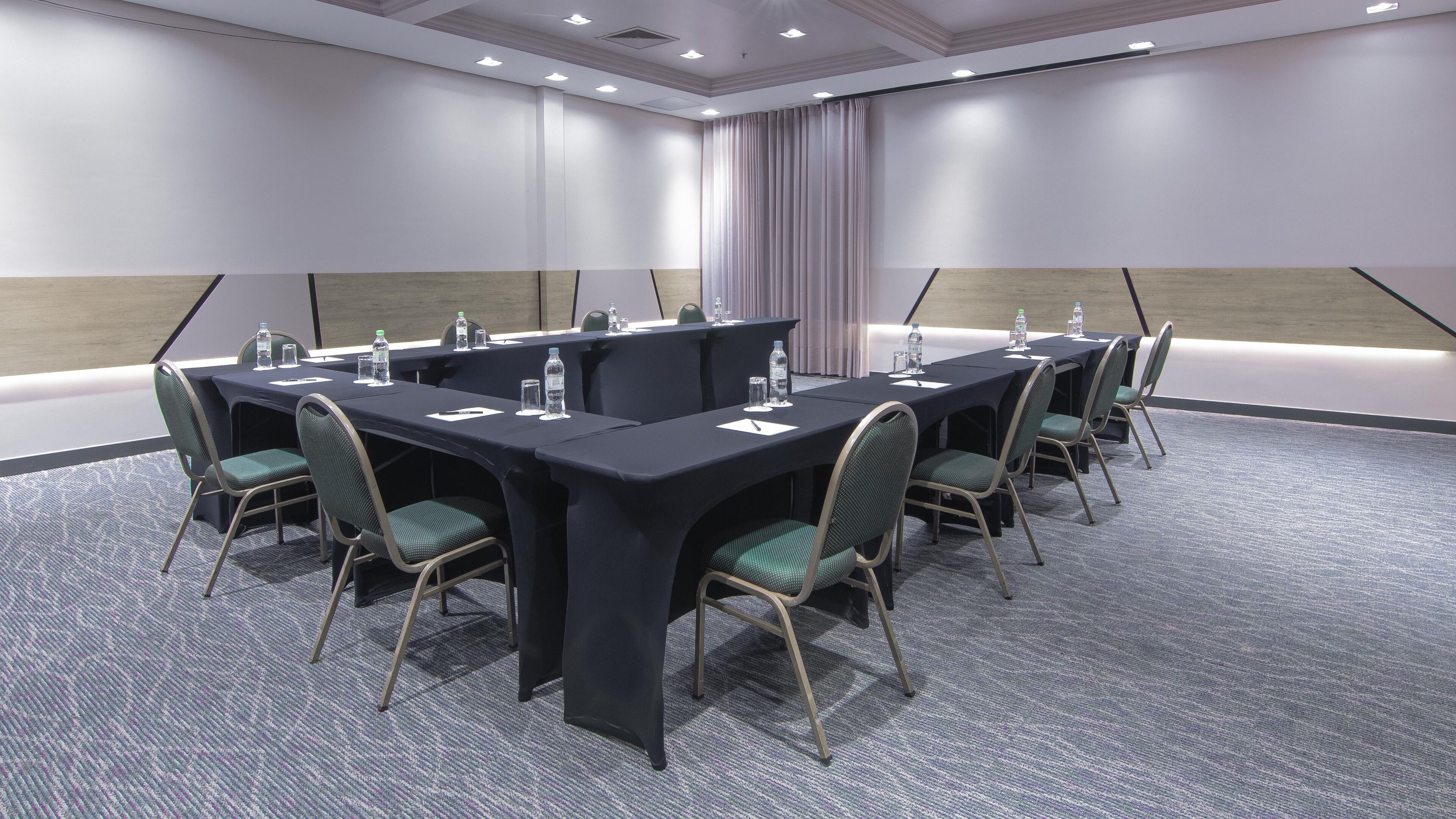 Uma sala de conferências com uma mesa comprida, cadeiras, garrafas de água e uma cortina.