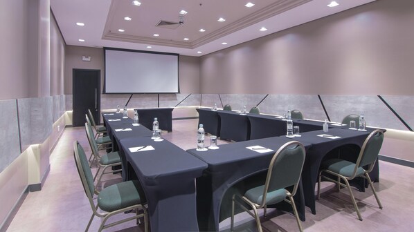 Une salle de conférence équipée de longues tables, de chaises, de bouteilles d’eau et d’un écran de projection.