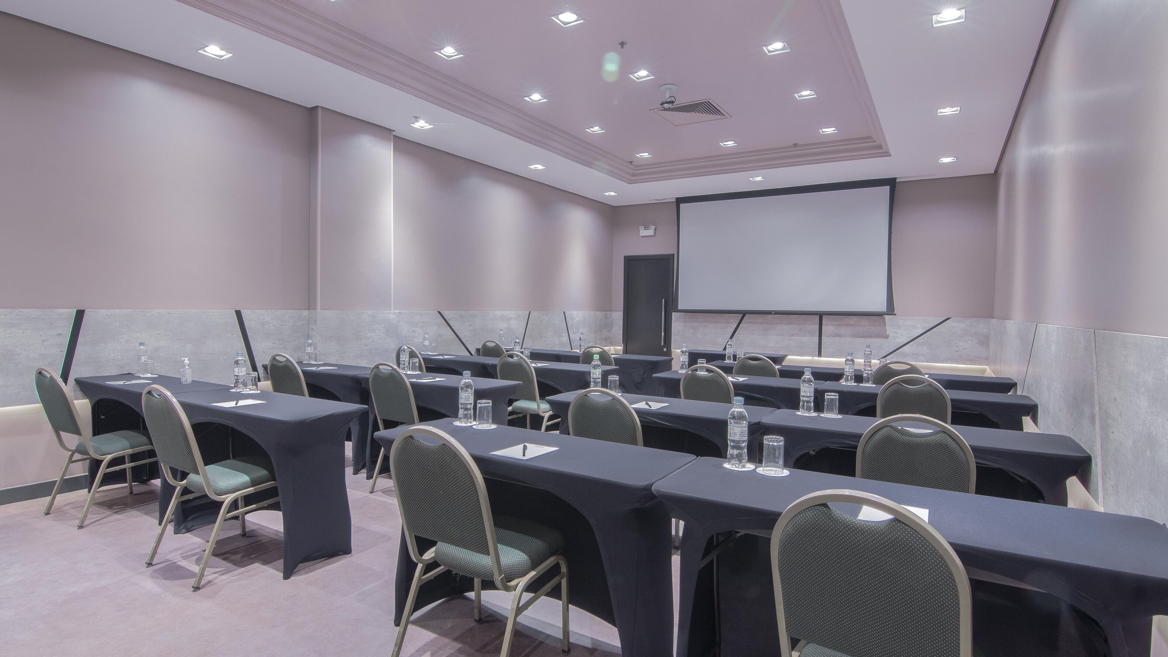 A conference room with multiple tables and chairs, equipped with water bottles and pens, and a projector screen.