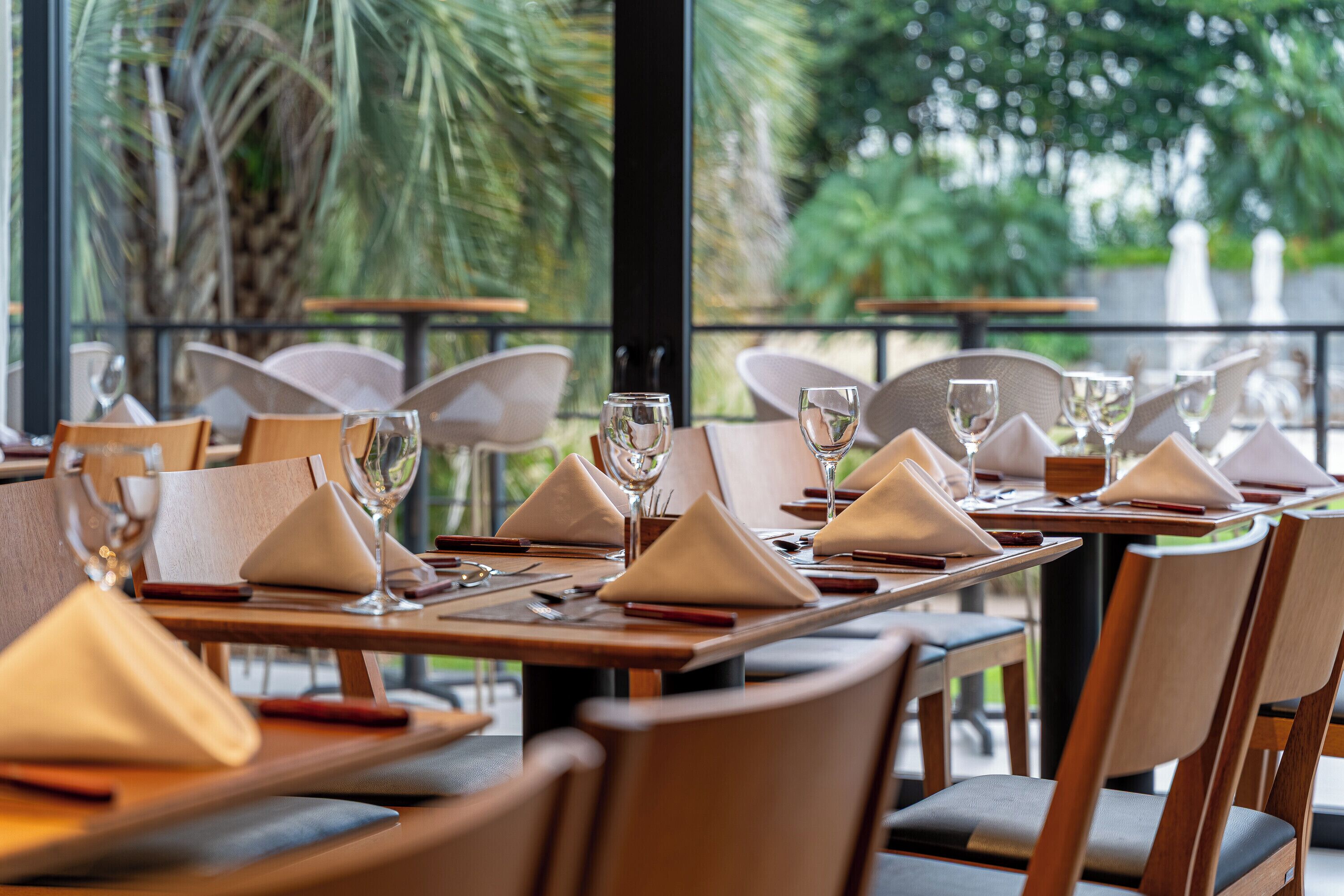 A restaurant setting with tables set for dining, chairs, and a view of greenery outside.
