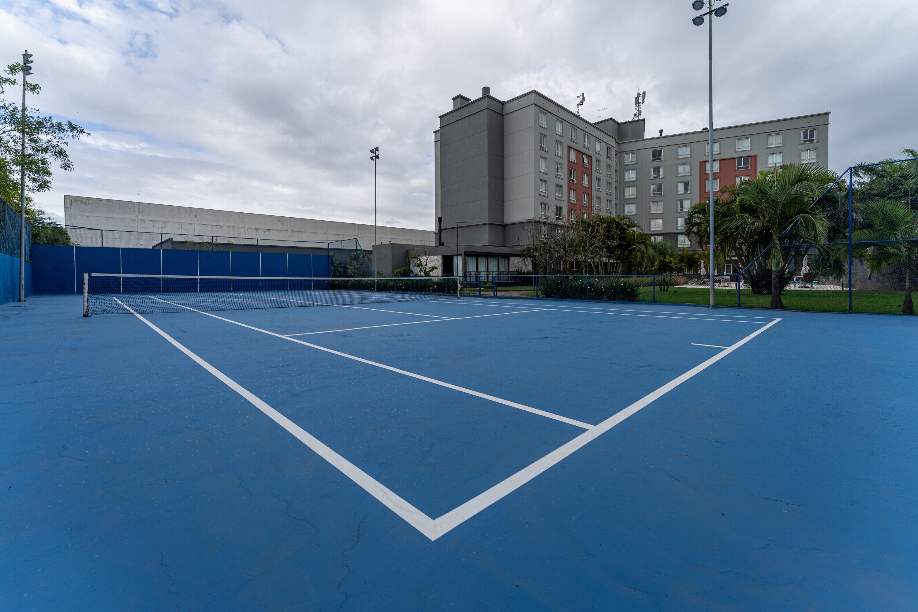 A tennis court with a net, surrounded by a blue fence and a building with multiple windows.