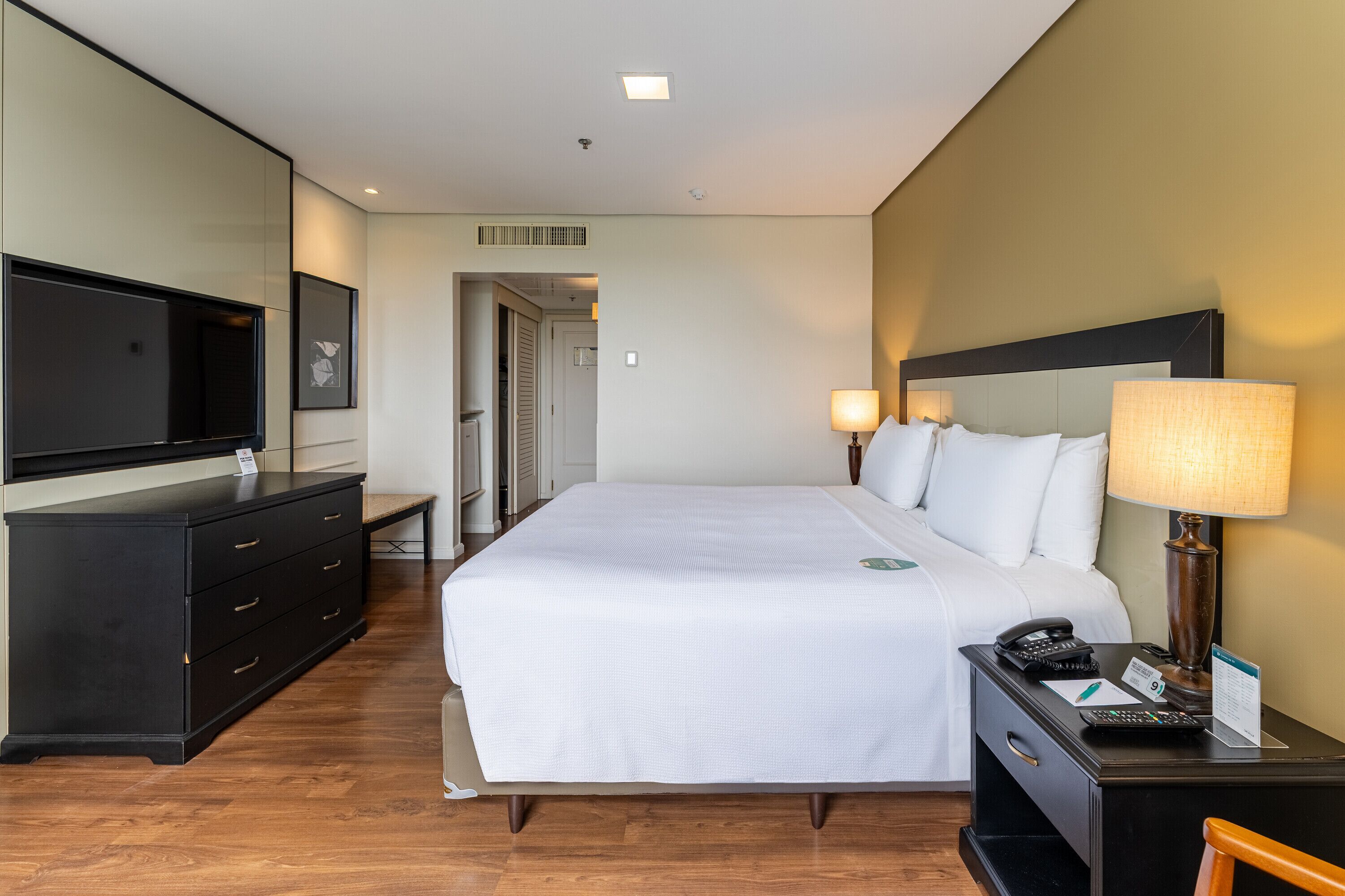 A hotel room with a bed, bedside table, lamp, and a television on a dresser.