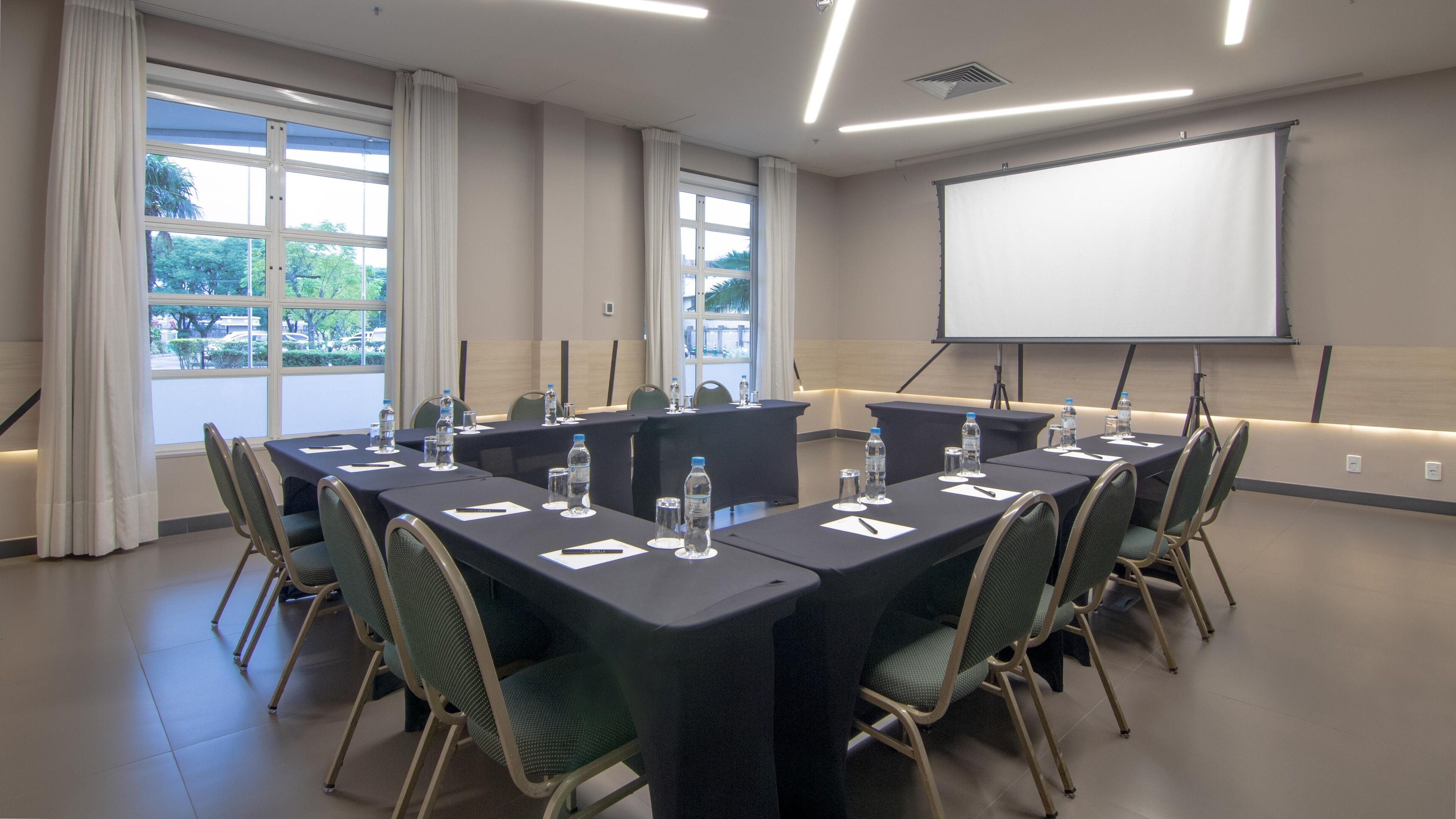 Un salón de conferencias con una gran mesa, sillas, pantalla de proyector y ventanas con cortinas.
