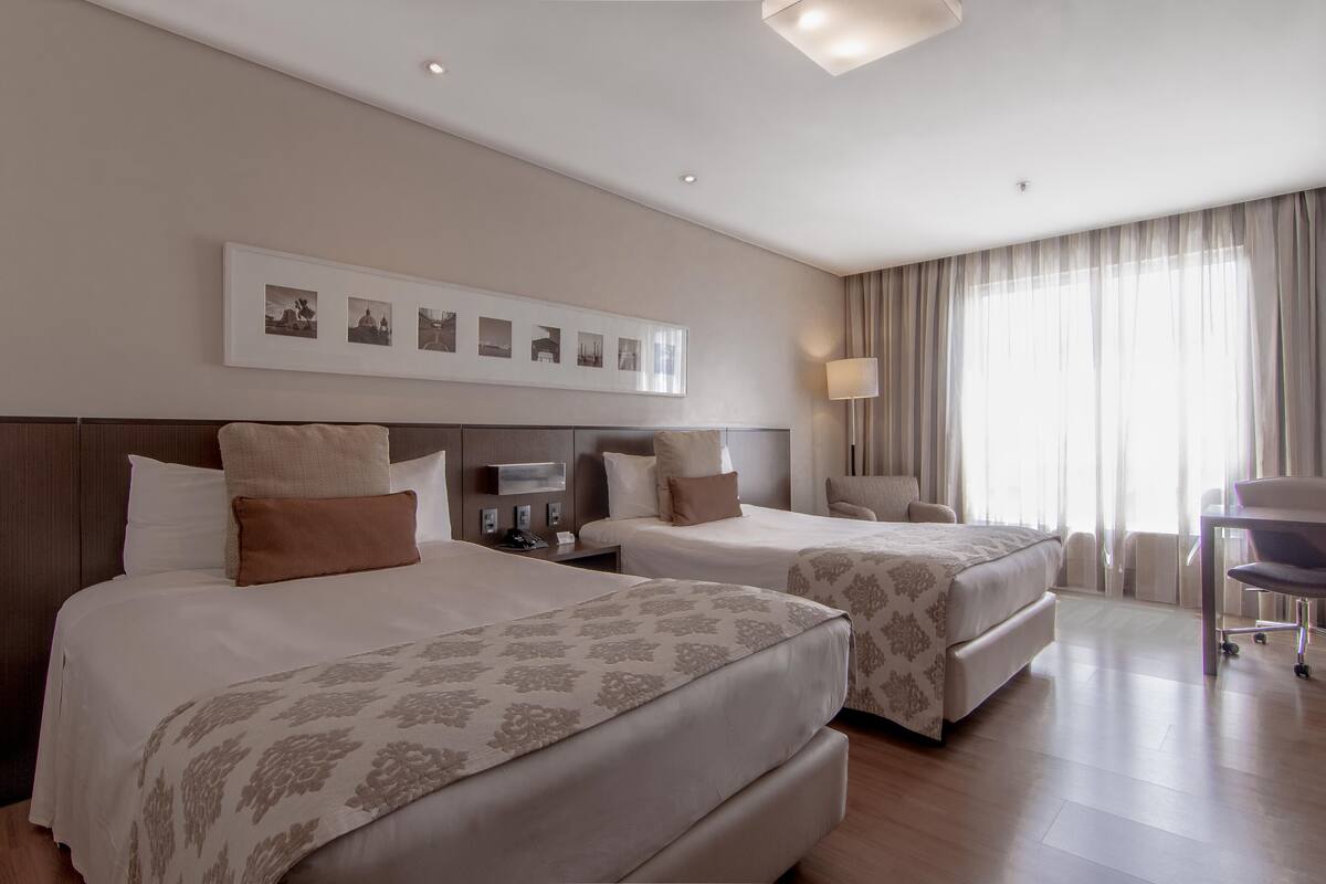 Een hotelkamer met twee bedden, een bureau, een stoel en een groot raam met gordijnen.