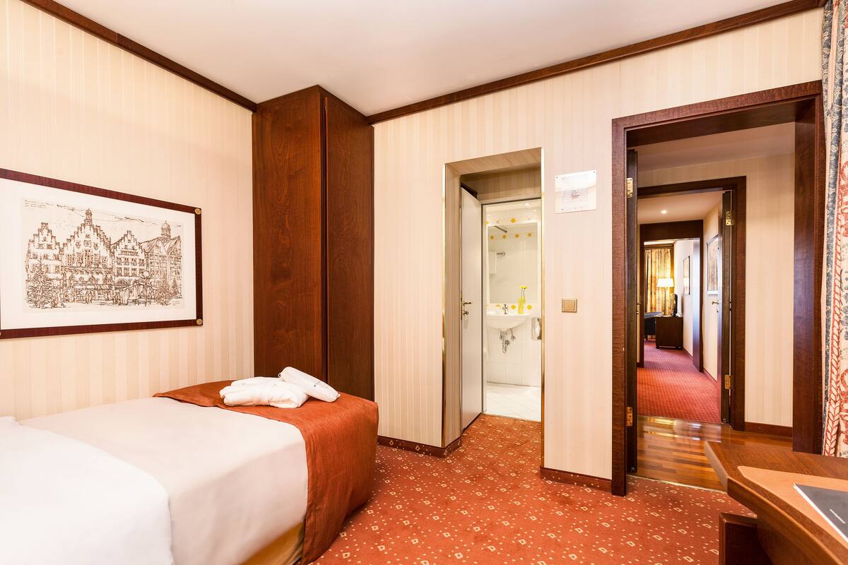 Habitación de hotel con cama, armario de madera, baño visible a través de una puerta abierta y un cuadro en la pared.