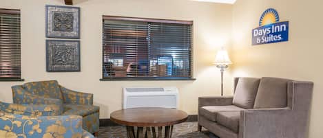 Una habitación de hotel con un sofá azul floral, un sillón gris, una mesa de centro de madera y una lámpara. Hay un ventanal con persianas, un calentador de pared y un cartel de Days Inn & Suites.