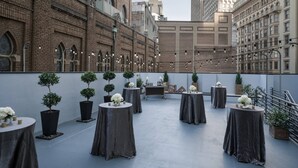 Una terraza en la azotea con mesas redondas, plantas en macetas y un edificio de ladrillo al fondo.
