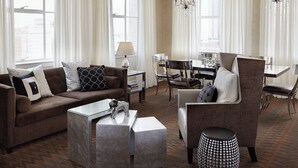 Una sala moderna con un sofá marrón, una mesa de centro de vidrio y un sillón estampado. Se observa un área de comedor con una mesa y sillas al fondo.
