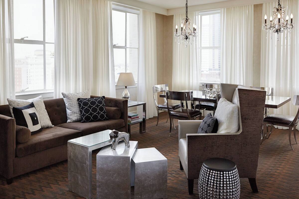 Een moderne woonkamer met een bruine bank, een glazen salontafel en een fauteuil met een patroon. Achtergrond toont een eethoek met een tafel en stoelen.