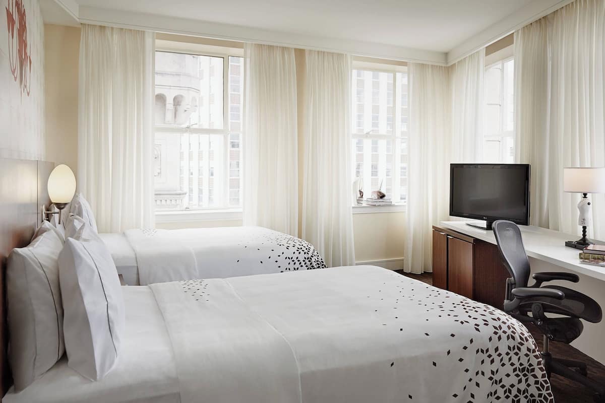 Een hotelkamer met twee bedden, een bureau met een televisie en grote ramen met gordijnen.