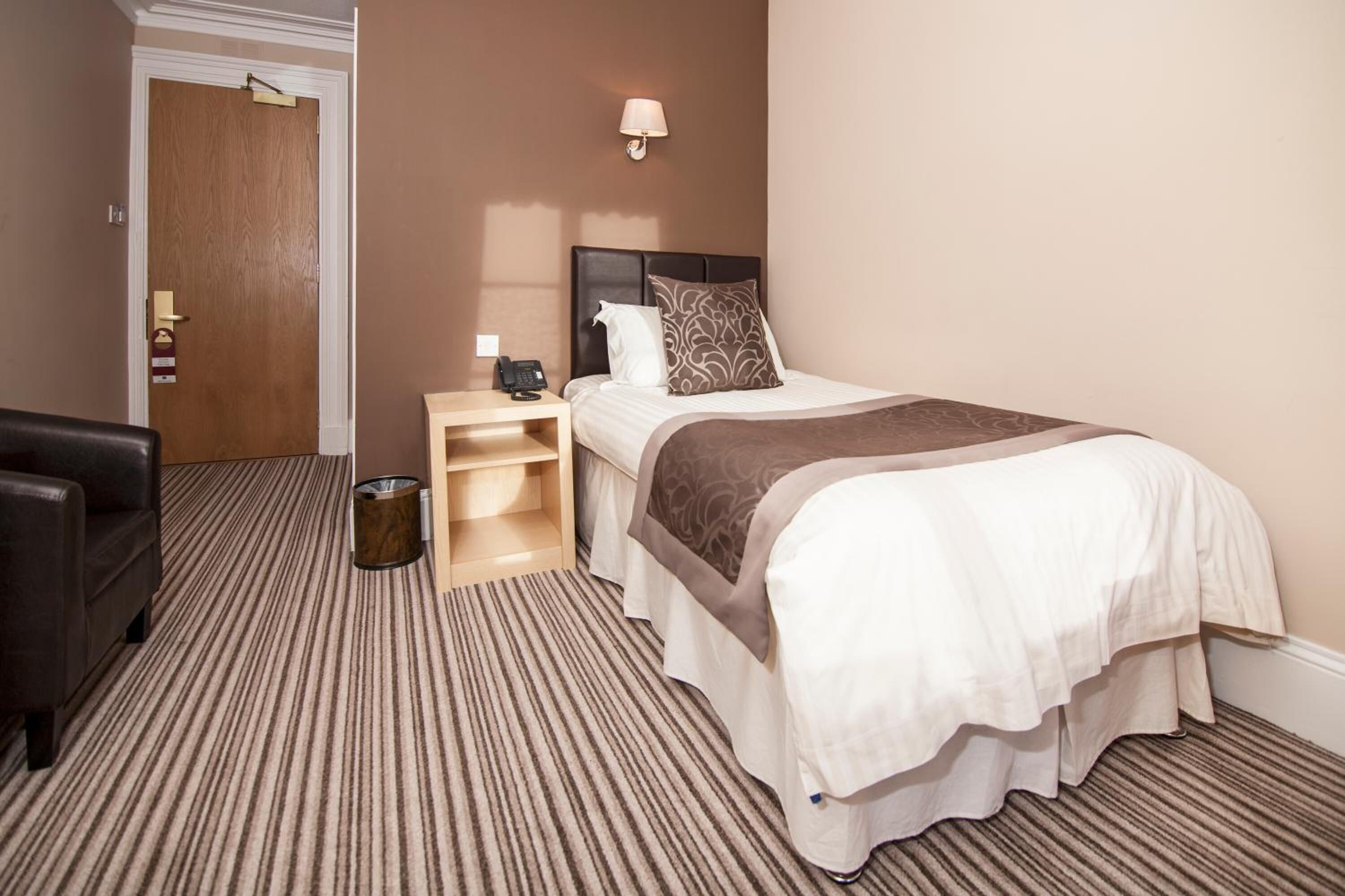 A hotel room with a bed, a bedside table, a lamp, a trash bin, and a door.